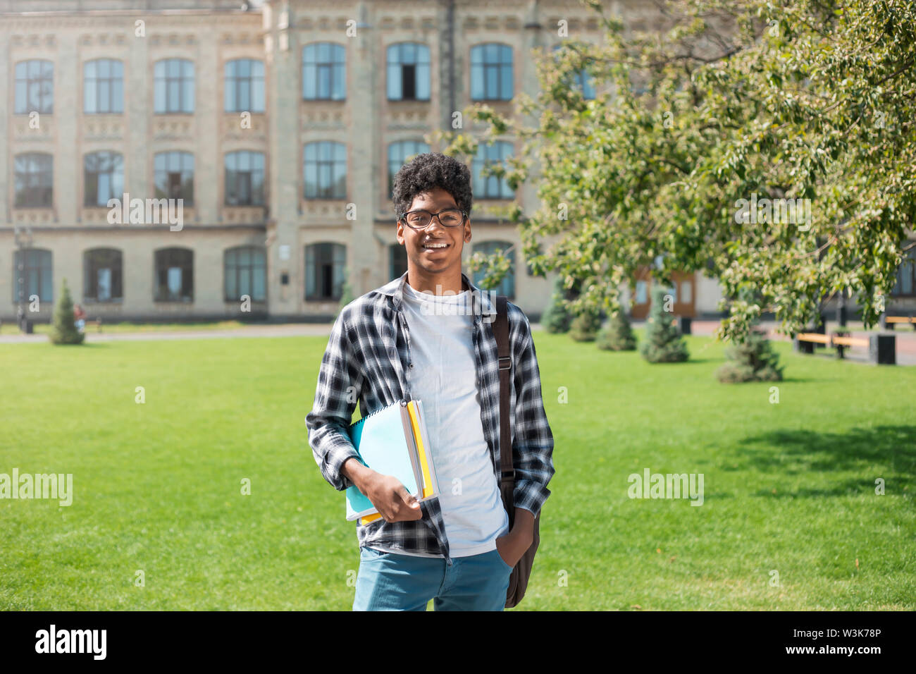 Un étudiant africain souriant avec des lunettes et des livres près de l'université. Portrait d'un jeune homme noir heureux debout sur un fond universitaire. Banque D'Images