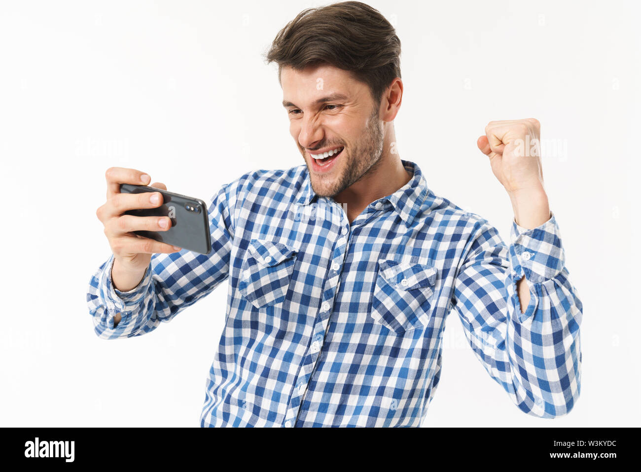 Photo de grand homme en chemise décontractée jouer jeu vidéo sur cellulaire et un poing serrant isolé sur fond blanc Banque D'Images