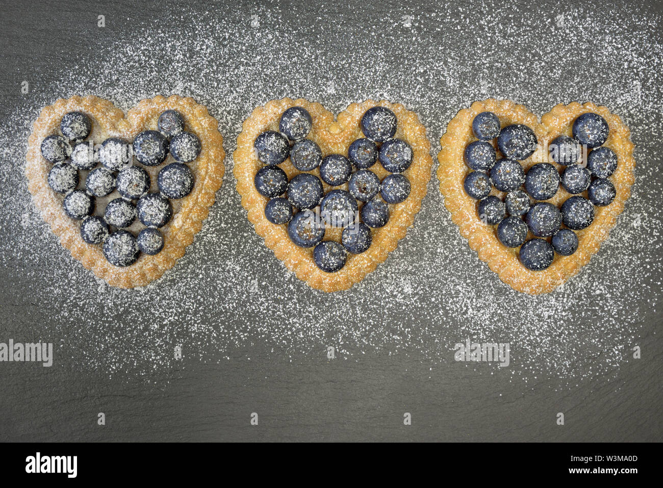 Trois petit gâteau en forme de coeur avec des bleuets se situent sur une plaque en ardoise noire et sont saupoudrés de sucre en poudre Banque D'Images