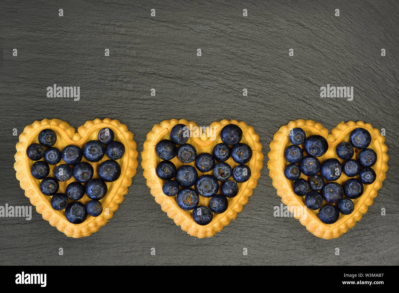 Trois petits gâteaux en forme de coeur avec des bleuets se situent sur une plaque en ardoise noire Banque D'Images