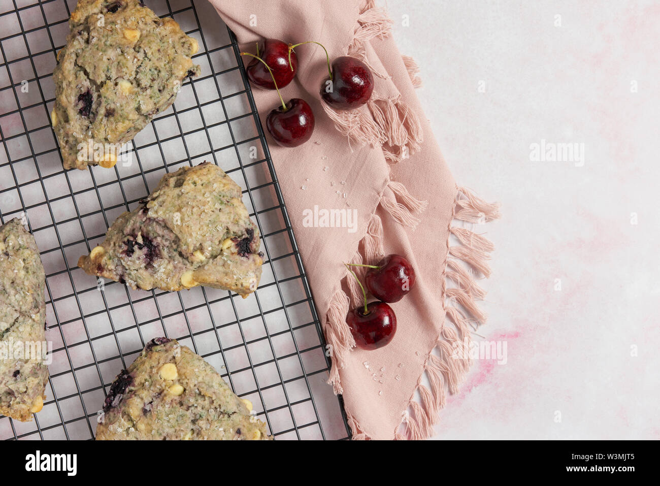 Scones au chocolat blanc et cerise noire sur une grille de cuisson et rose en arrière-plan avec une serviette et cerises Banque D'Images
