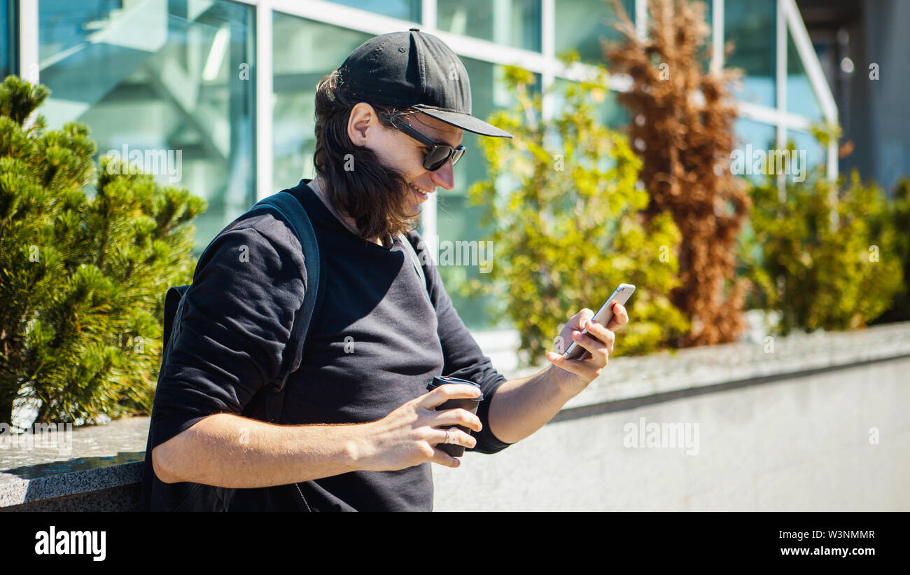 Un homme à lunettes noires, une casquette noire et des pousses de café sur la rue, sur un smartphone. Vue de côté. Paparazzi. Enquêtes privées. Banque D'Images