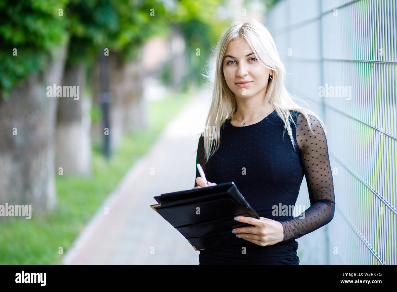 Une belle jeune fille blonde portant une robe noire classique est titulaire d'un dossier de documents dans ses mains. Style de vêtements d'affaires. La jeune fille travaille Banque D'Images