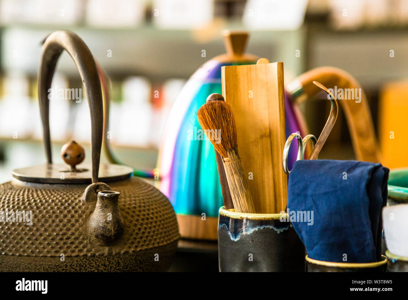 Une théière en fonte tetsubin se tient à côté d'une bouilloire laquée irisée multicolore et d'ustensiles pour la cérémonie du thé japonaise à la boutique de thé ANMO à Düsseldorf, en Allemagne. Une cuillère à thé en bambou (chashaku) et un fouet à thé (chasen) reposent dans une tasse en céramique sombre. Banque D'Images