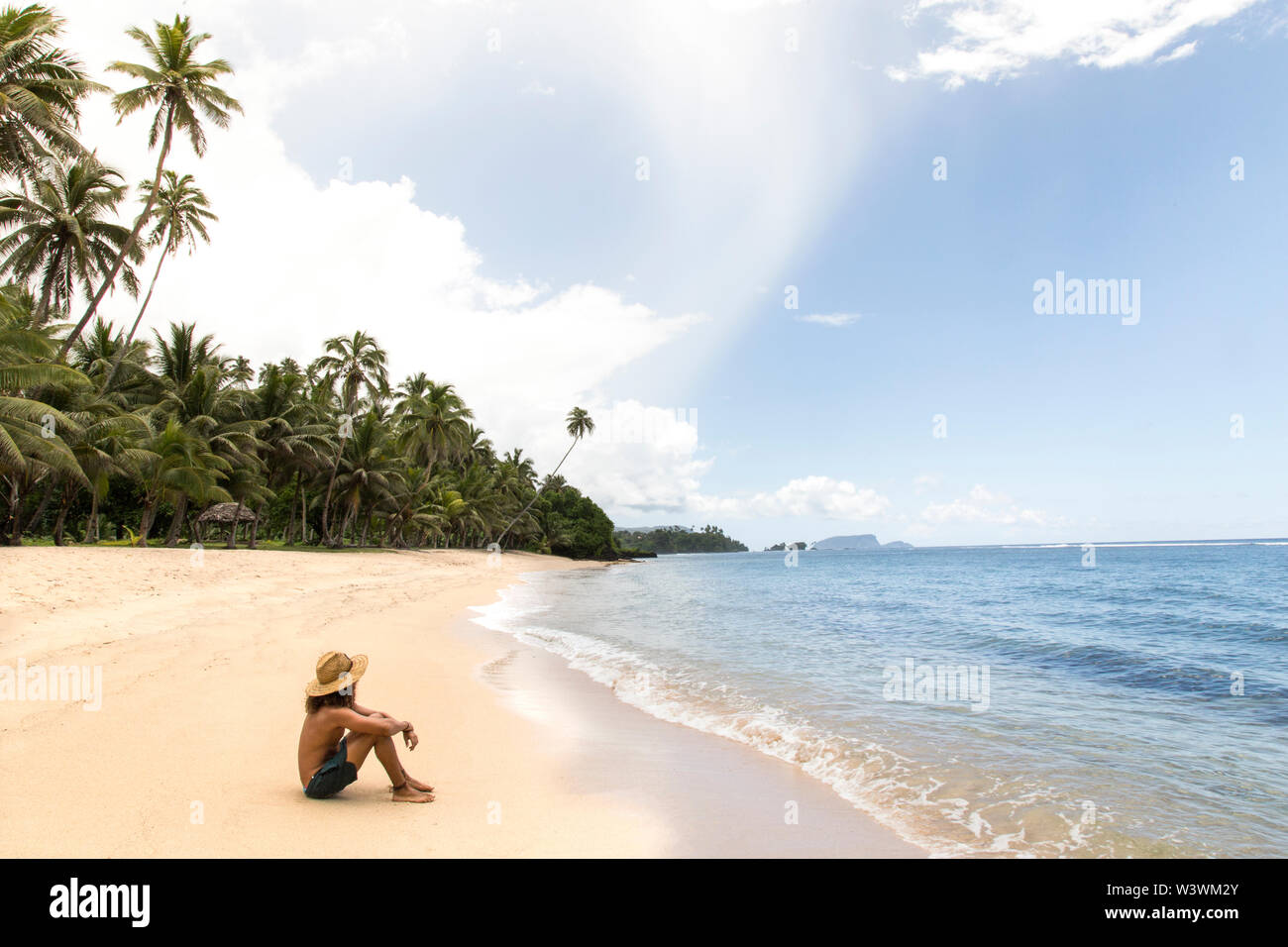 Un homme, wearing hat, détente à la plage tropicale de sable de Samoa Banque D'Images