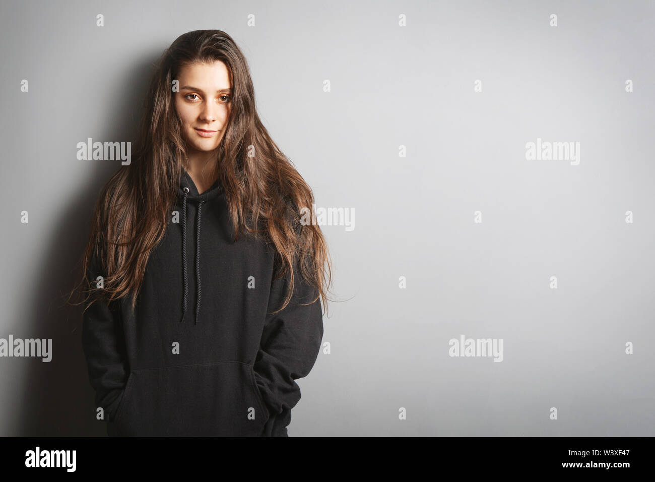 Cool jeune femme avec de longs cheveux de brunette wearing black hoodie sweater leaning against wall avec ses mains dans les poches - fond gris with copy space Banque D'Images