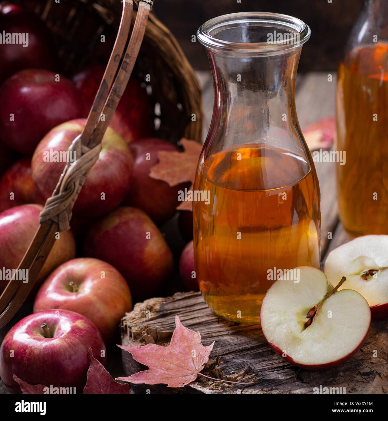 Pichet de cidre et pomme coupé en deux sur bois rustique avec un panier de pommes rouges renversé en arrière-plan Banque D'Images