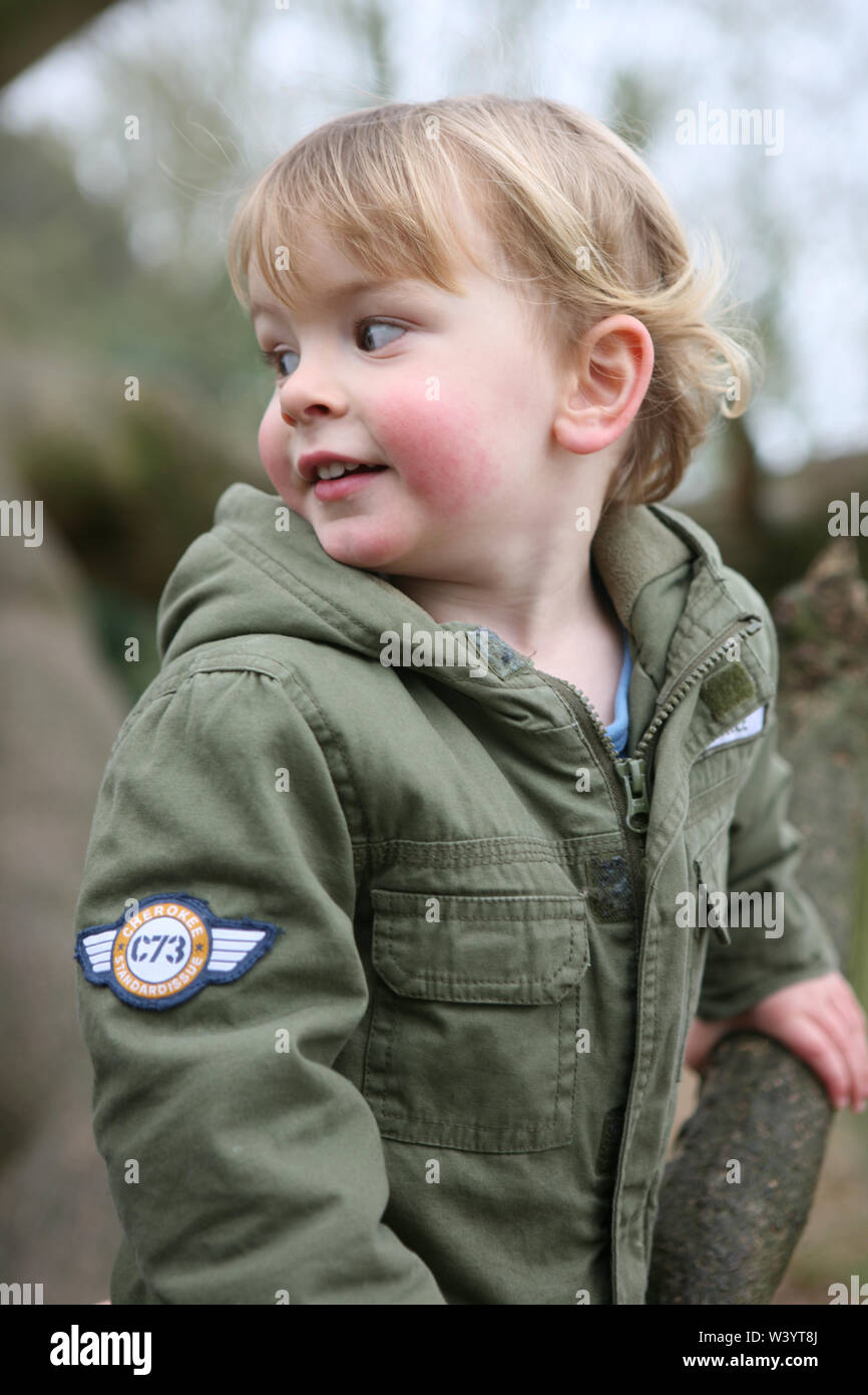 Petit garçon (2/3 ans) portant un manteau kaki, souriant et regardant par-dessus son épaule : Mottisfont, Hampshire, Royaume-Uni. Parution du modèle Banque D'Images