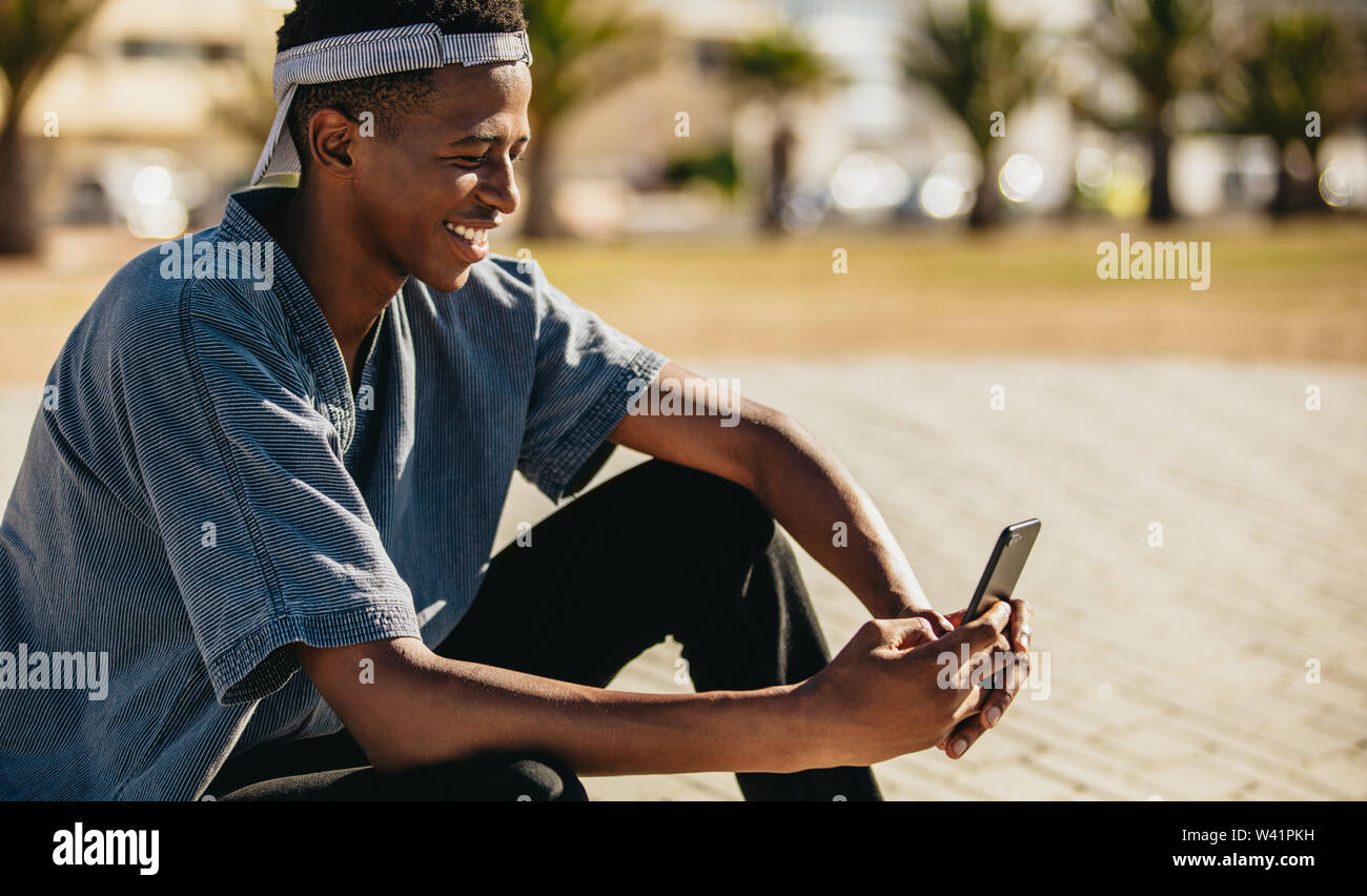 African American jeune mec assis à l'extérieur et à l'aide d'un téléphone cellulaire. Young man texting avec son téléphone mobile et de sourire, Banque D'Images