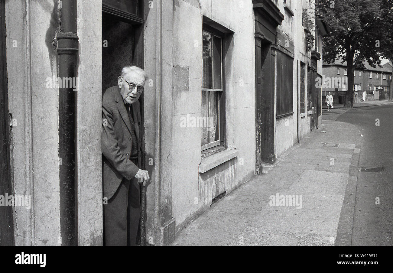 Début des années 1970, historique, un frêle vieillard dans une veste et cravate avec bâton de marche debout à la porte de son ancienne maison victorienne, aperçu dans Charlton village, dans le sud de Londres, Angleterre, Banque D'Images