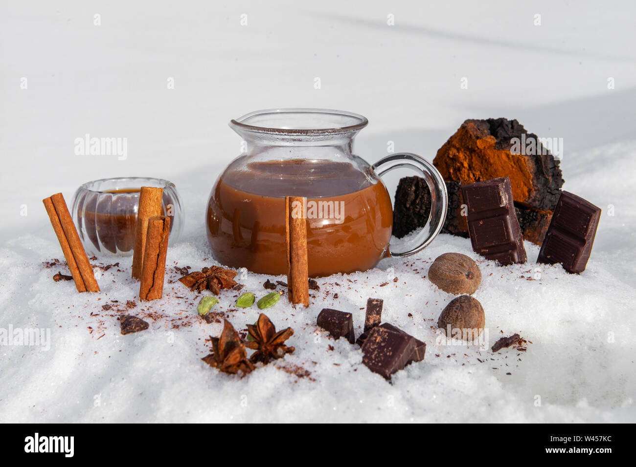 Les bâtons de cannelle, les herbes et épices sont vus de près, debout dans la neige par une urne en verre rempli de thé chai chaga, une ancienne recette utilisée dans la médecine alternative Banque D'Images