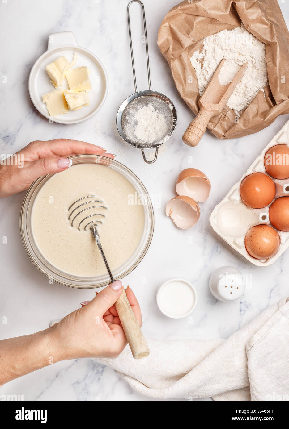 Femme prépare la pâte à crêpes maison pour le petit-déjeuner. Mélanger au fouet pour fouetter dans les mains. Ingrédients sur la table - farine de blé, œufs, beurre, sucre, sa Banque D'Images
