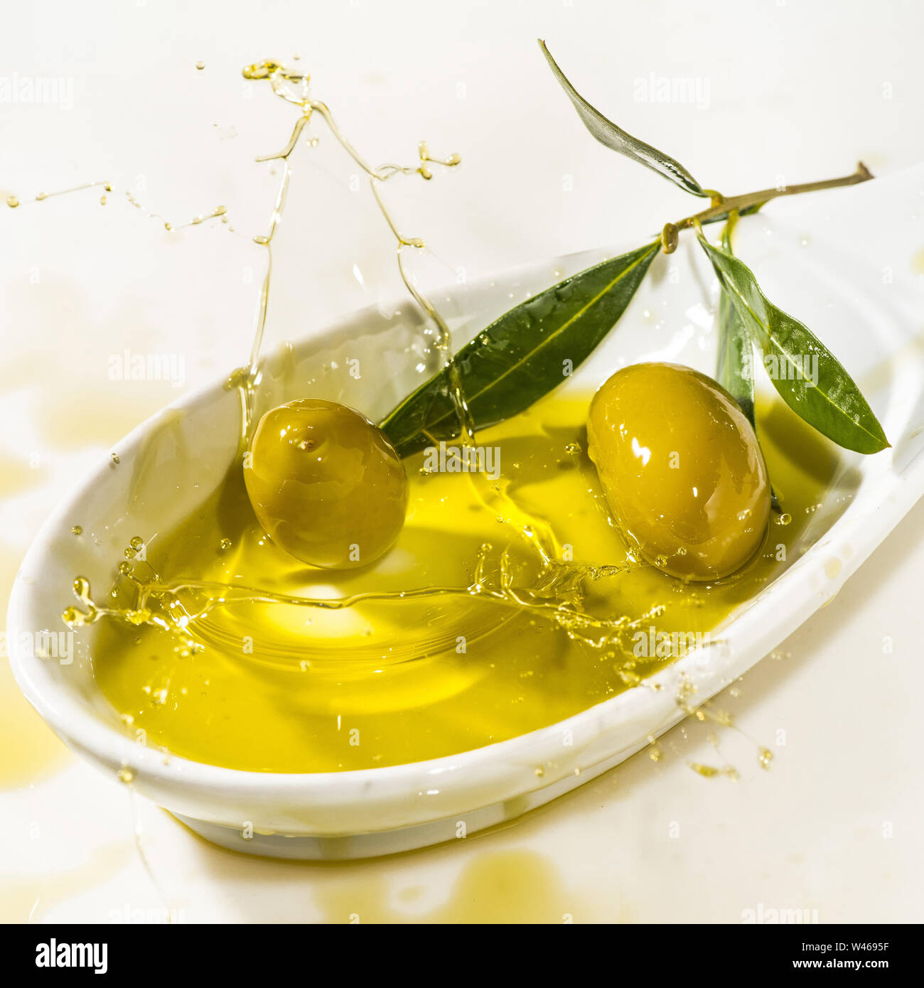 Quelques olives vertes tombent dans un bol rempli d'huile d'olive avec splash. Fond blanc Banque D'Images