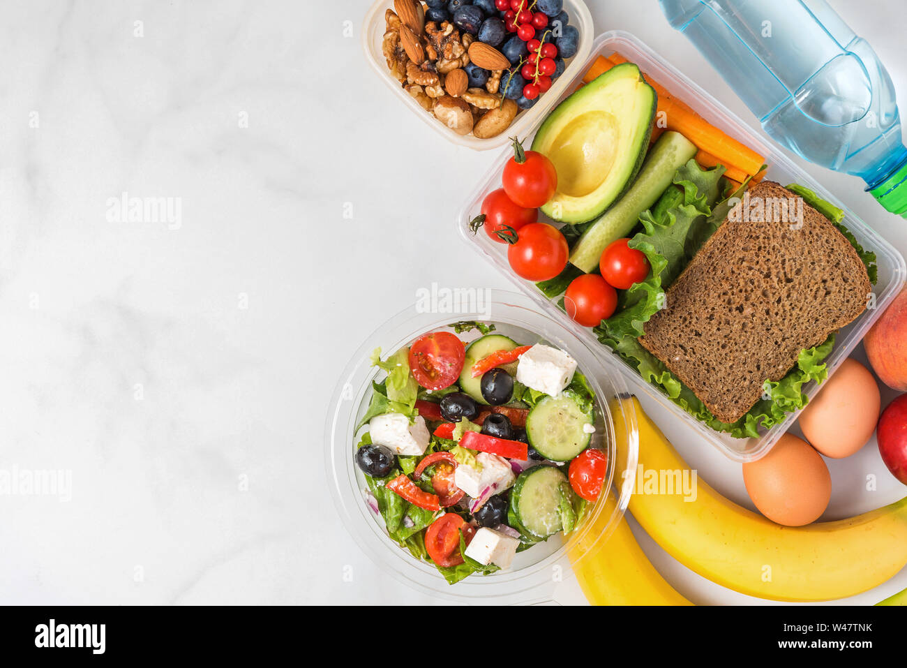 Boîtes à lunch santé avec sandwich, salade grecque, des légumes, des noix, des baies, des bananes, des œufs, de l'avocat, des pêches et de l'eau sur fond blanc. Vue de dessus avec Banque D'Images