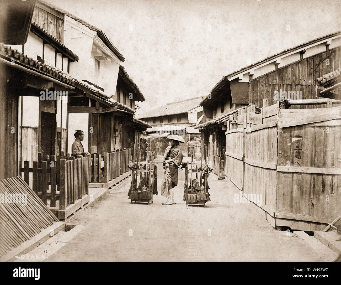 [ 1870 ] Backstreet Japon - Osaka - un balai vendeur parle avec un homme debout en face d'une porte dans une rue d'Osaka, ca 1870. Il n'y a pas de poteaux électriques et lampes à gaz encore. La première d'Osaka lampe à essence a été inaugurée en 1871 (4) l'ère Meiji, alors que l'électricité a été introduite vers 1887 (20) L'ère Meiji. 19e siècle vintage albumen photo. Banque D'Images