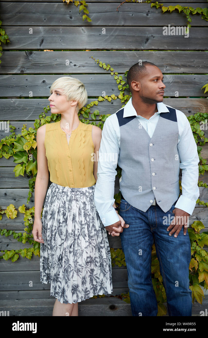 Élégant et attrayant multicultural couple amoureux se tenant la main par une clôture dans un cadre urbain rempli de lierre Banque D'Images