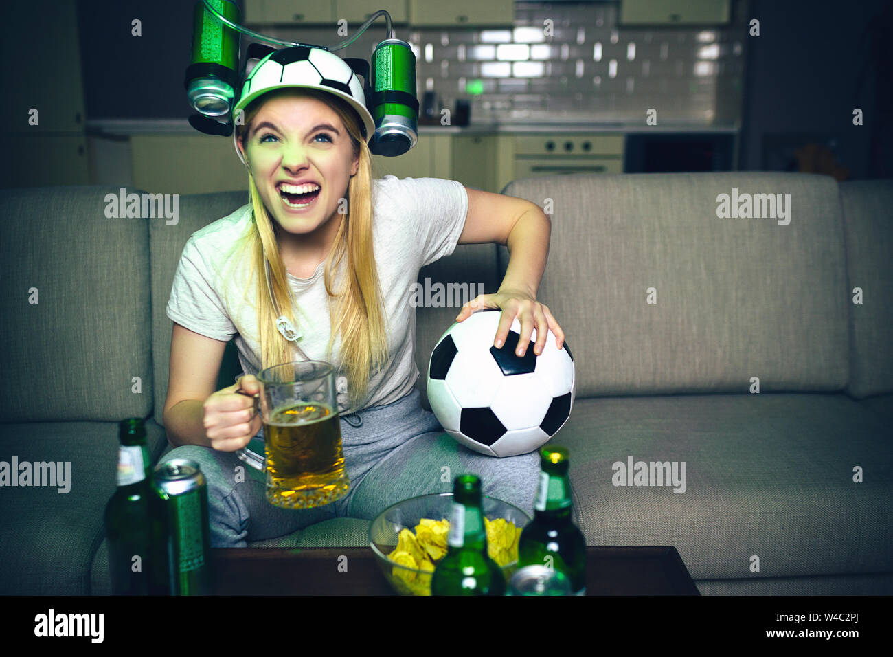 Jeune femme regarder match de football à la télévision le soir. Modèle en colère s'asseoir sur un canapé et maintenir le verre de bière avec le ballon. Cheering seul. Chapeau spécial avec des canettes de bière o Banque D'Images