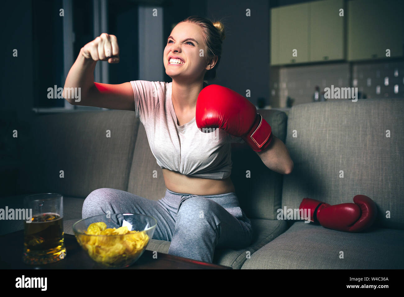 Jeune femme watch boxe sur plat dans la nuit. Modèle émotionnel en colère. Cheering seul. Porter des gants de boxe rouge. La malbouffe sur table. Les combats à la fin Banque D'Images