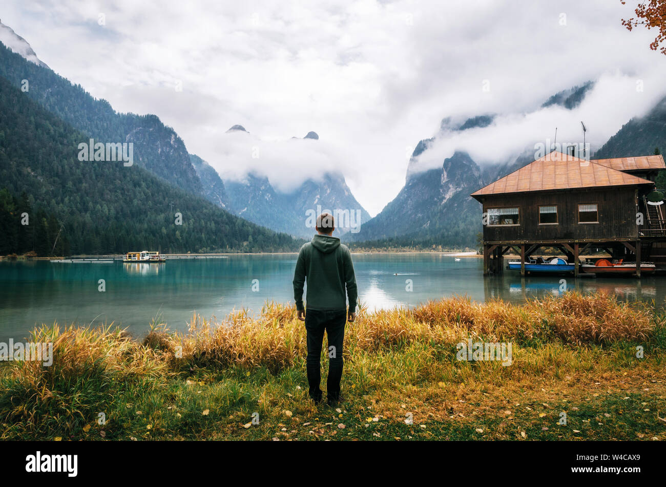 Jeune homme hiker est dos à la caméra sur la rive du lac Dobbiaco ou Toblacher Dolomites dans un hangar à bateaux en bois avec cabane, Italie Banque D'Images