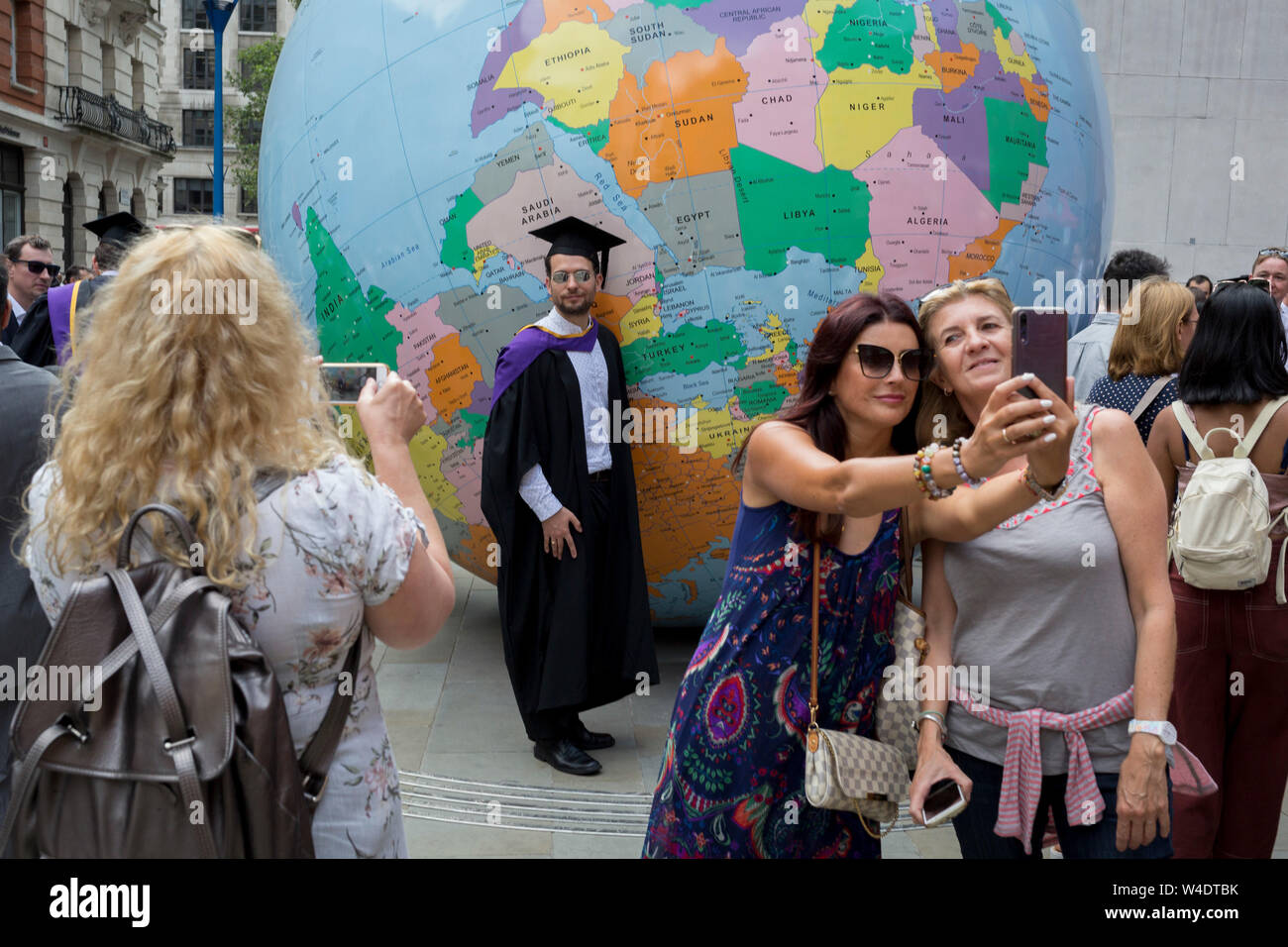 Sous la sculpture de l'artiste lauréat du Prix Turner Mark Wallinger intitulée "Le Monde à l'envers', les nouveaux diplômés directement après leur cérémonie de fin rencontrez la famille et les amis à l'extérieur de la London School of Economics (LSE), le 22 juillet 2019, à Londres, en Angleterre. "Le Monde à l'envers" est un grand globe politique, quatre mètres de diamètre, avec des États nations et frontières décrites mais avec la simple et révolutionnaire twist d'être inversée. La plupart des masses continentales se trouvent maintenant dans le "bas" avec les pays de l'hémisphère et les villes ré-étiquetés pour cette nouvelle orientation. Banque D'Images