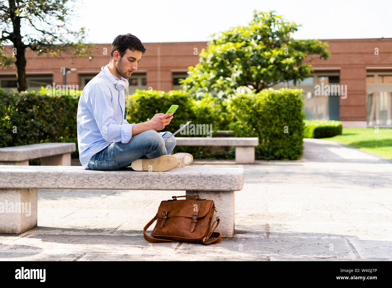 Jeune homme à l'aide d'ordinateur portable et le smartphone, écouteurs autour du cou, assis sur un banc Banque D'Images