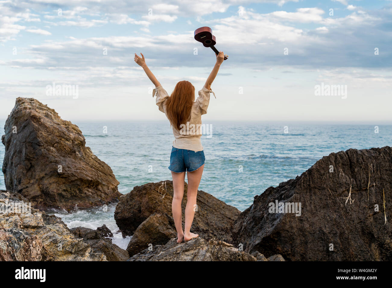 Vue arrière de jeune femme avec ukulele debout sur la roche face à la mer Banque D'Images