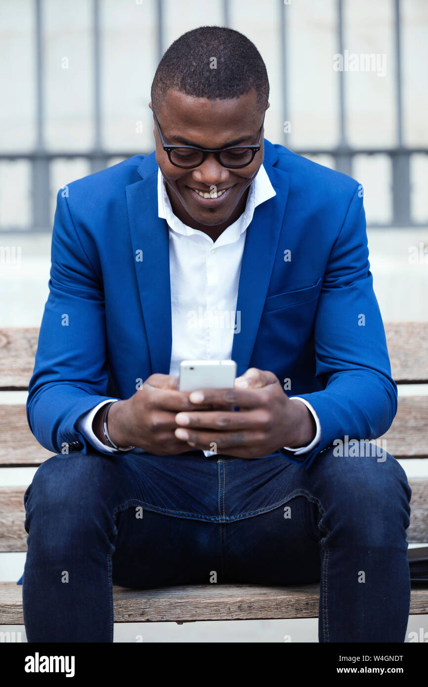 Young businessman wearing blue veste de costume assis sur un banc et using smartphone Banque D'Images