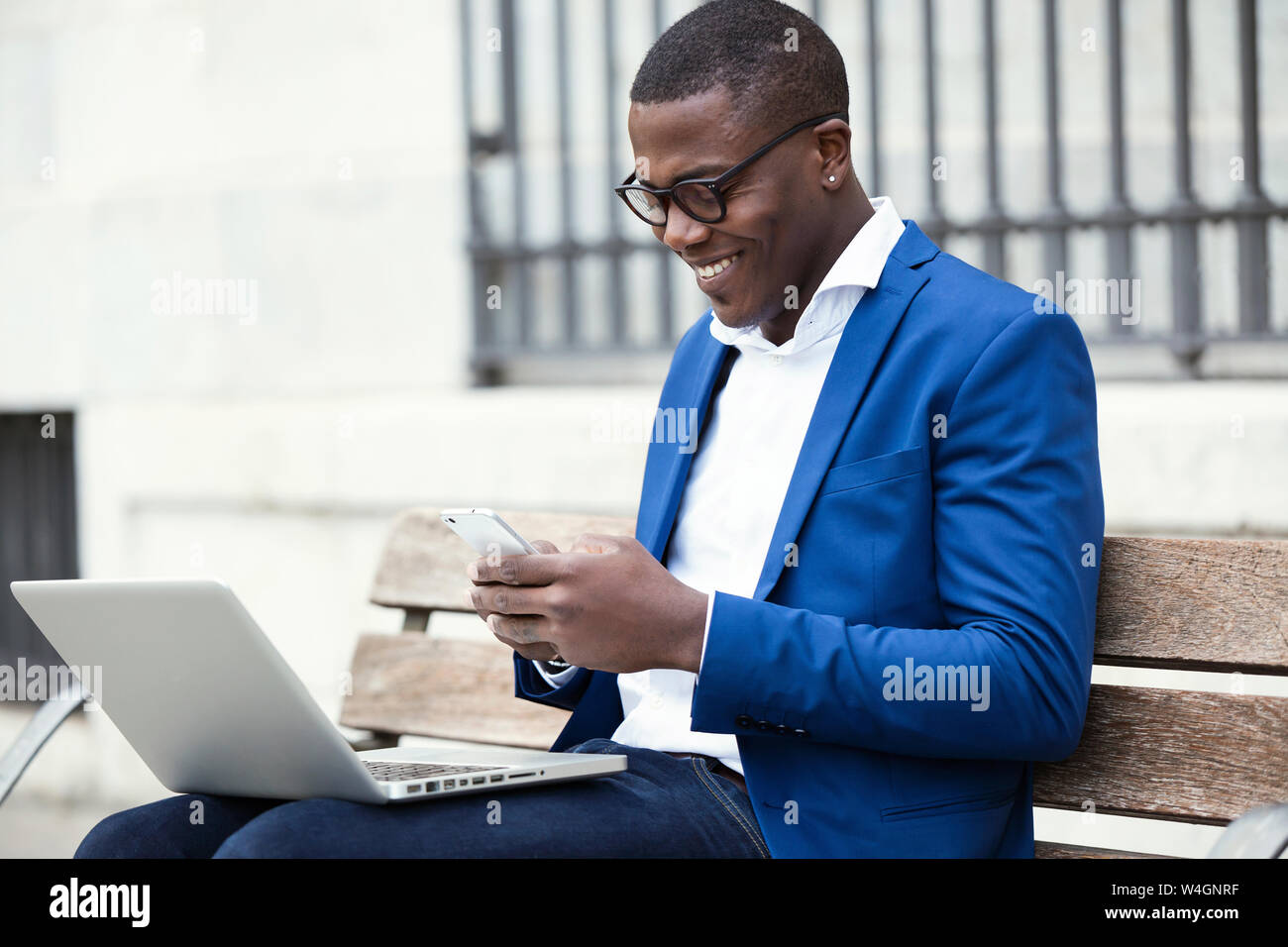 Young businessman wearing blue veste de costume assis sur un banc et using smartphone Banque D'Images
