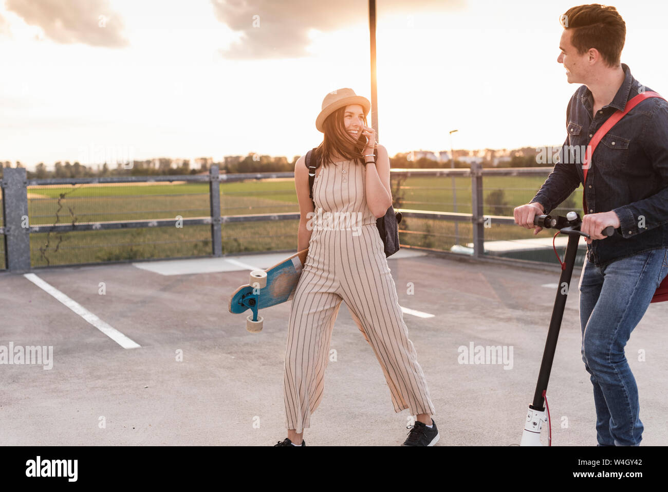 Happy young couple with cell phone, longboard et scooter électrique sur parking Banque D'Images