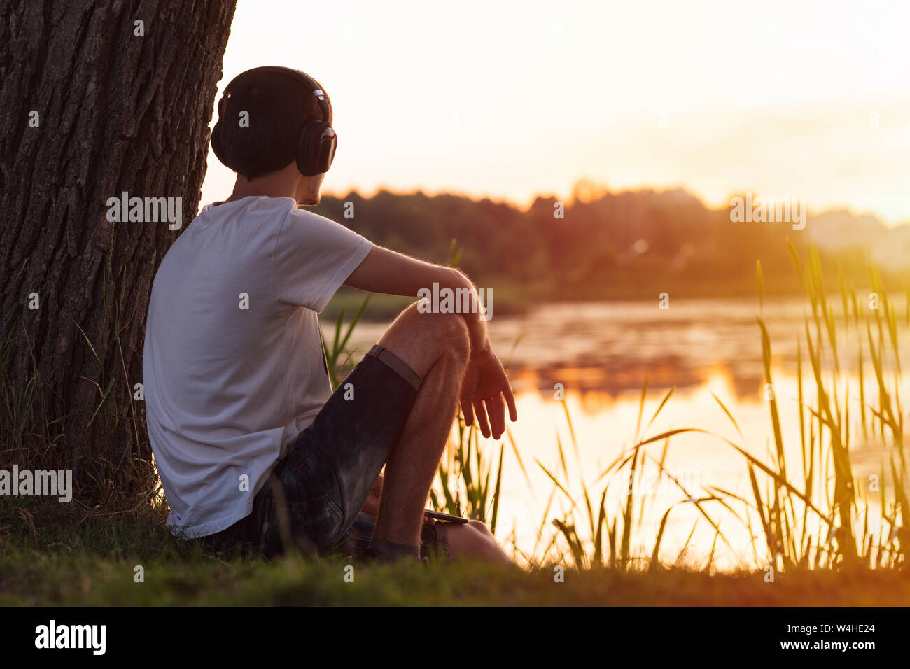 Jeune homme solitaire assis sur le lac par un arbre au coucher du soleil et d'écouter de la musique avec des écouteurs. Un week-end de détente dans la campagne ou le parc. Banque D'Images