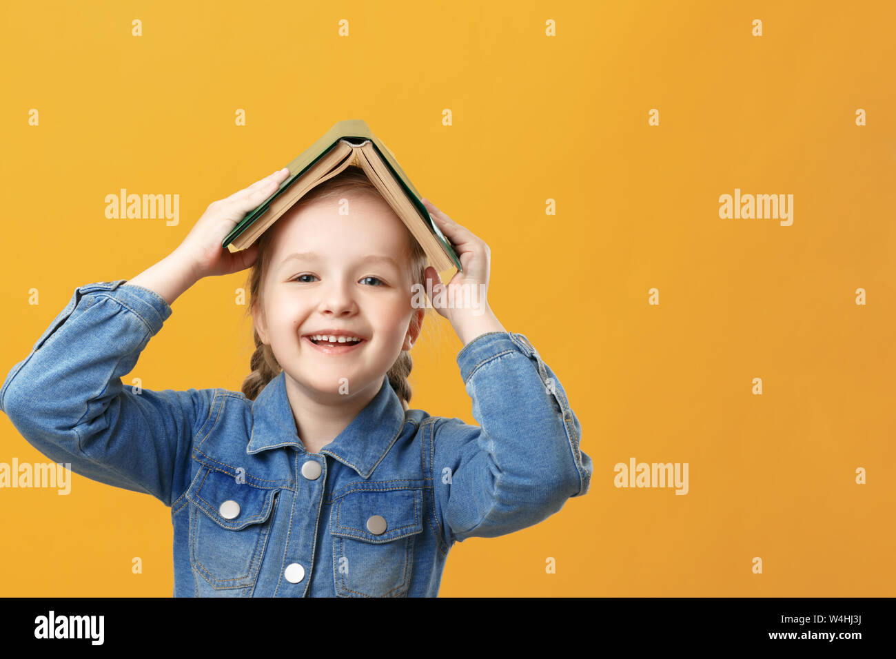 Portrait d'une drôle de petite fille sur un fond jaune. L'élève a un livre sur la tête. Banque D'Images