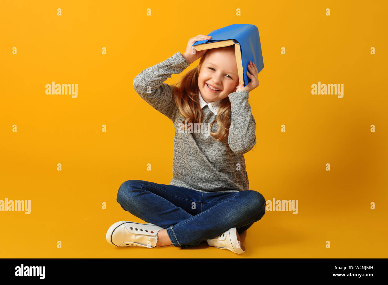 Jolie petite fille joyeuse étudiant est assis sur le plancher avec un livre sur la tête. Le concept d'éducation et de l'école. Fond jaune. Studi Banque D'Images