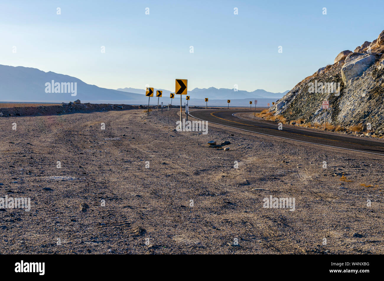 Desert route tournant autour de paroi rocheuse avec ciel voilé au-delà des montagnes du désert. La signalisation routière indiquant une route en courbe. Banque D'Images