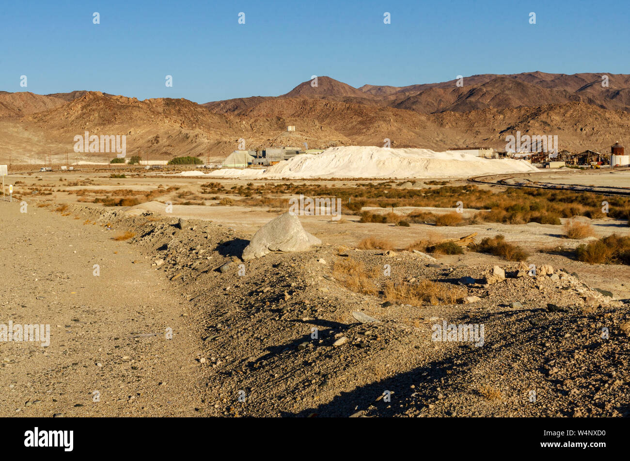 Route de terre menant vers les montagnes du désert aride sous un ciel bleu, sable blanc comme des piles de dire et la ville au loin. Banque D'Images