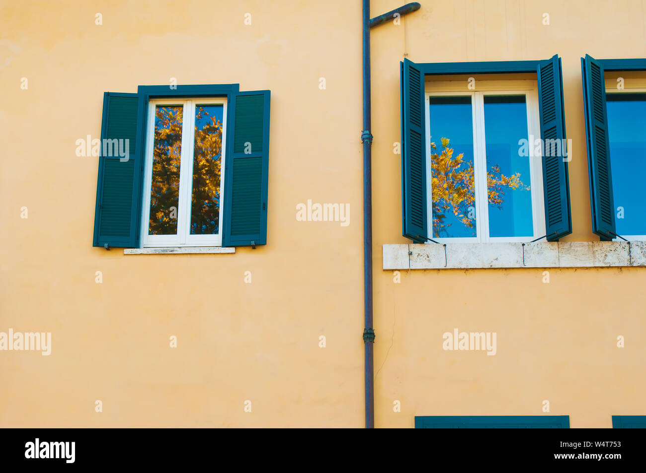 Deux fenêtres avec volets vert foncé sur un mur jaune uni. Reflet de ciel bleu et jaune des branches d'arbre. Rome, Italie Banque D'Images