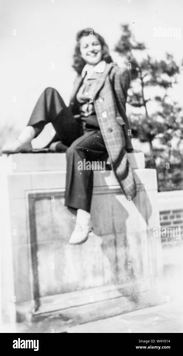 Full Length portrait of young woman smiling, assis à l'extérieur sur un mur de béton et de briques, 1950. () Banque D'Images