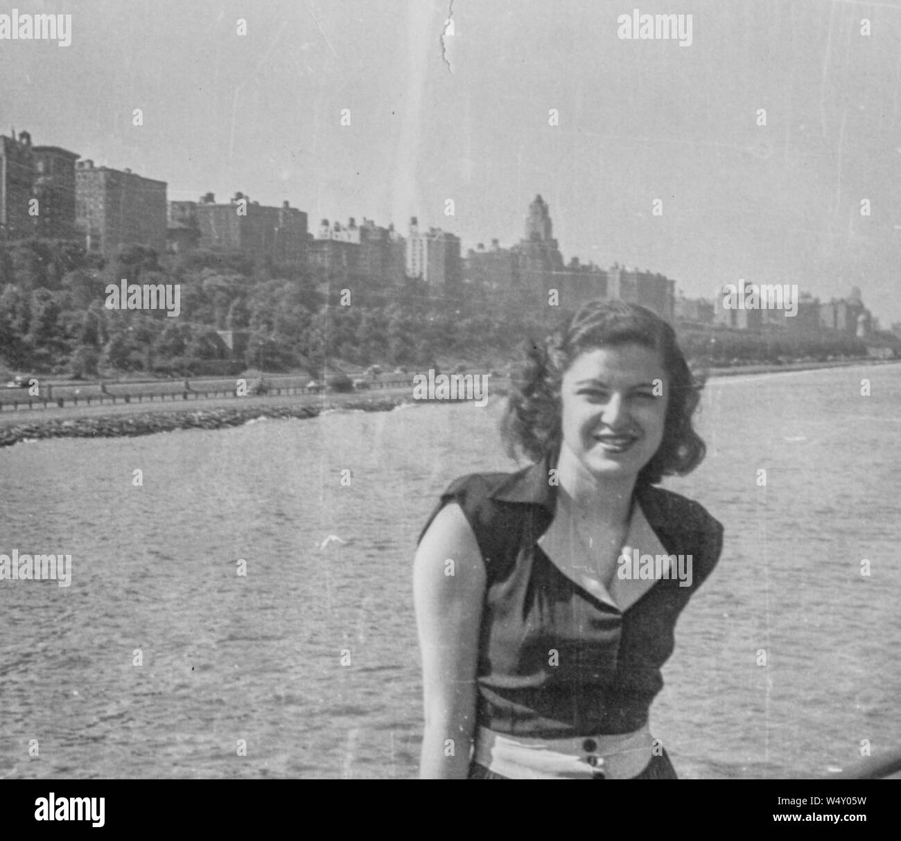 Demi-longueur portrait of young woman wearing dress assis sur la rampe d'un bateau et souriant, avec le paysage urbain d'une ville en arrière-plan, 1945. () Banque D'Images