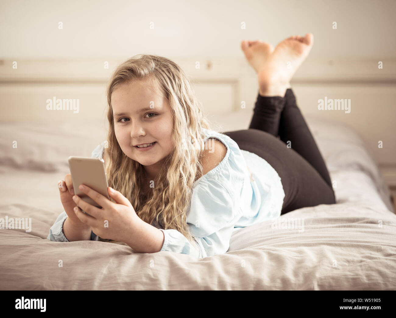 Belle blonde happy little girl playing sur téléphone mobile. Cute child discussion sur internet avec les médias sociaux smartphone app au lit à la maison. Banque D'Images