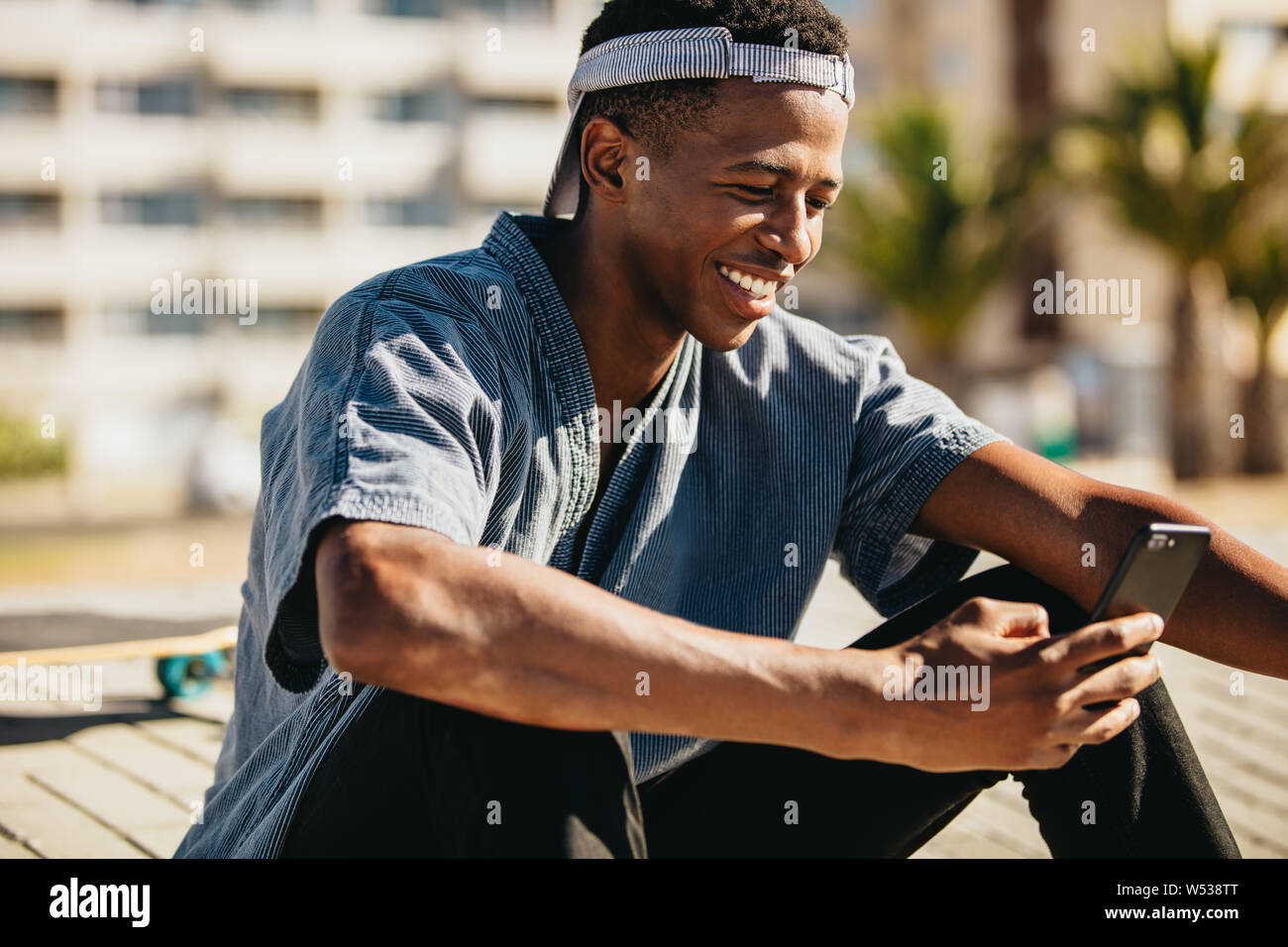 Smiling african american man sitting outdoors et la lecture des messages texte sur son téléphone intelligent. Jeune skateur professionnel smiling lors de l'utilisation de téléphone mobile. Banque D'Images