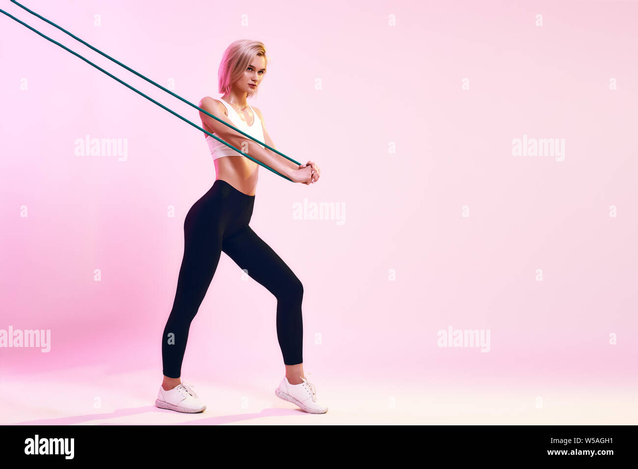 Organisme de formation . Vue latérale du jeune femme sportive en haut blanc et noir leggings exerçant avec bande de résistance en position debout contre fond rose en studio. Sport concept. L'équipement de sport Banque D'Images