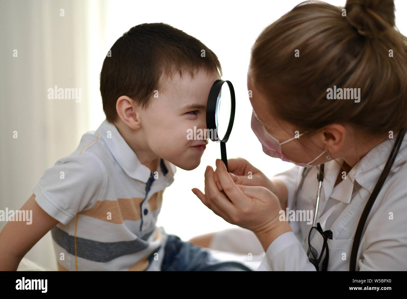 L'enfant fait une grimace et regarde le médecin à travers une loupe qu'une femme dans un manteau blanc tient dans ses mains. Banque D'Images