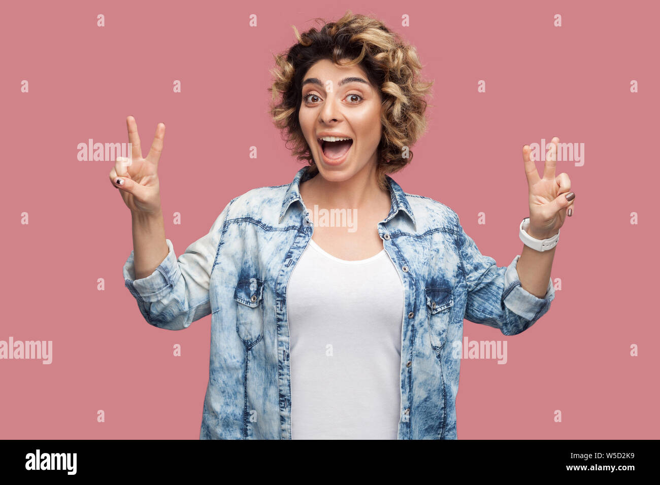 Portrait of happy funny jeune femme avec les cheveux bouclés chemise décontractée debout avec la victoire ou signe de la paix et des gestes à la caméra avec étonnement face à Banque D'Images