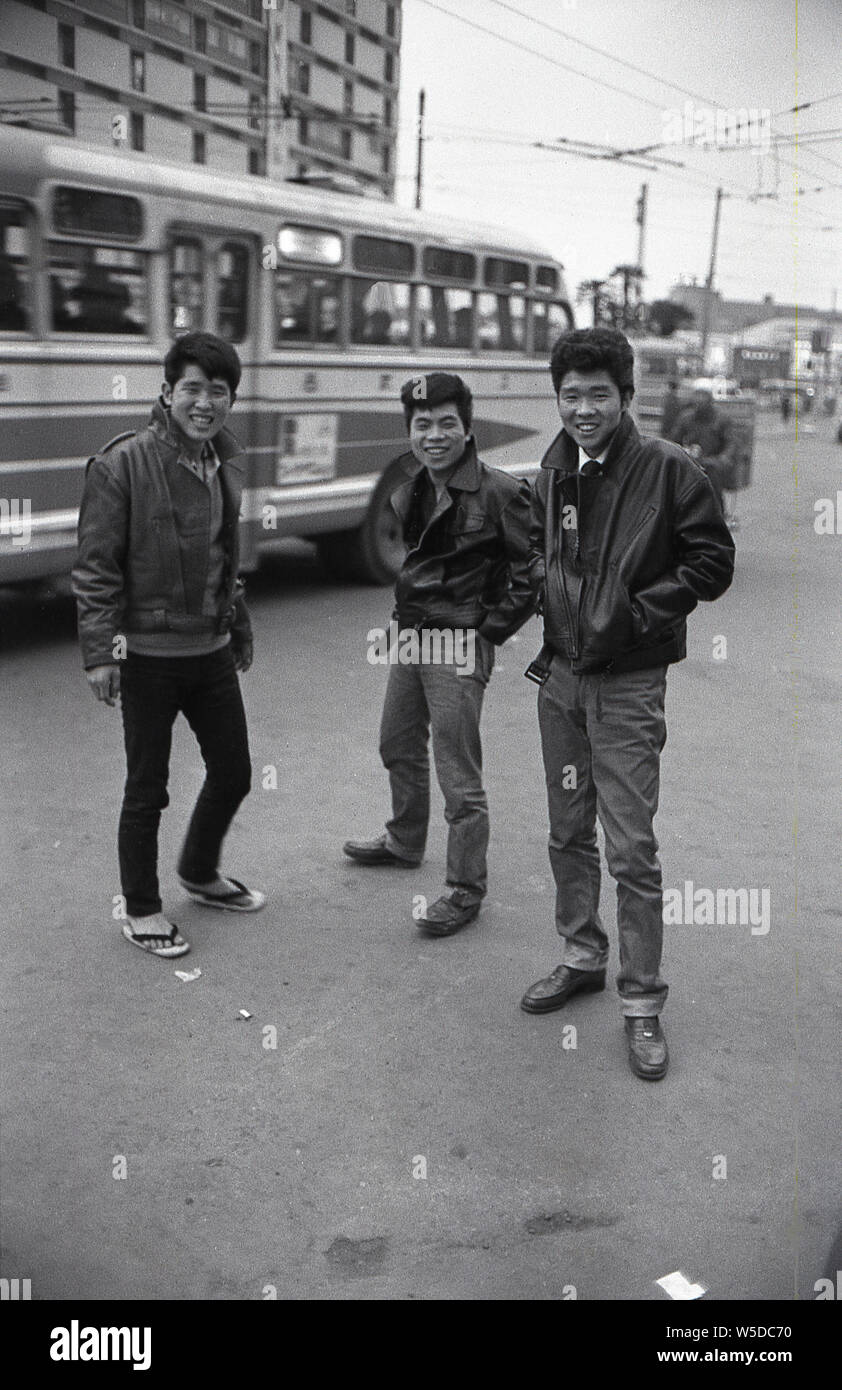 [ 1960 - Japon ] jeunes japonais à la mode - Trois jeunes hommes portant des coiffures à la mode japonaise, des jeans et des vestes en cuir, dans une imitation de l'apparence de graissage de l'Amérique, de se tenir sur une rue à Tokyo comme ville laissez-passer d'autobus, 1960 (Showa 35). Banque D'Images