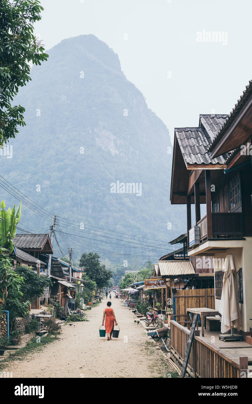 Muang Ngoi, Laos - Mai 2019 : femme laotienne en robe orange avec deux seaux de marcher sur la rue centrale du village Banque D'Images