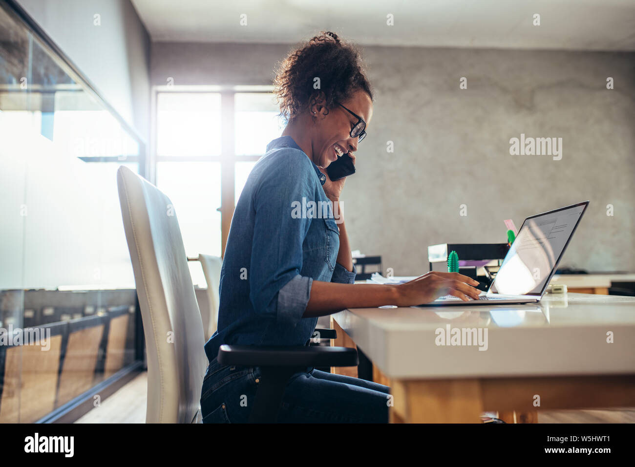 Vue latérale photo de femme parlant au téléphone et souriant tout en travaillant sur l'ordinateur portable. Businesswoman travaillant à son bureau. Banque D'Images