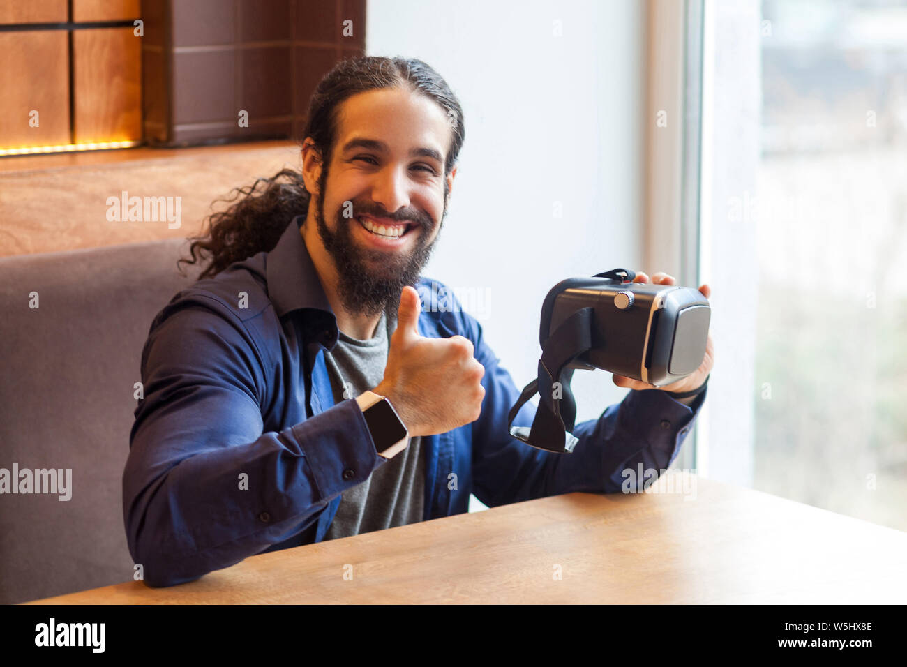 Portrait of happy young adult man barbu dans un style décontracté assis dans un café, et vr holding showing Thumbs up sur simulateur avec visage heureux, à la recherche en c Banque D'Images