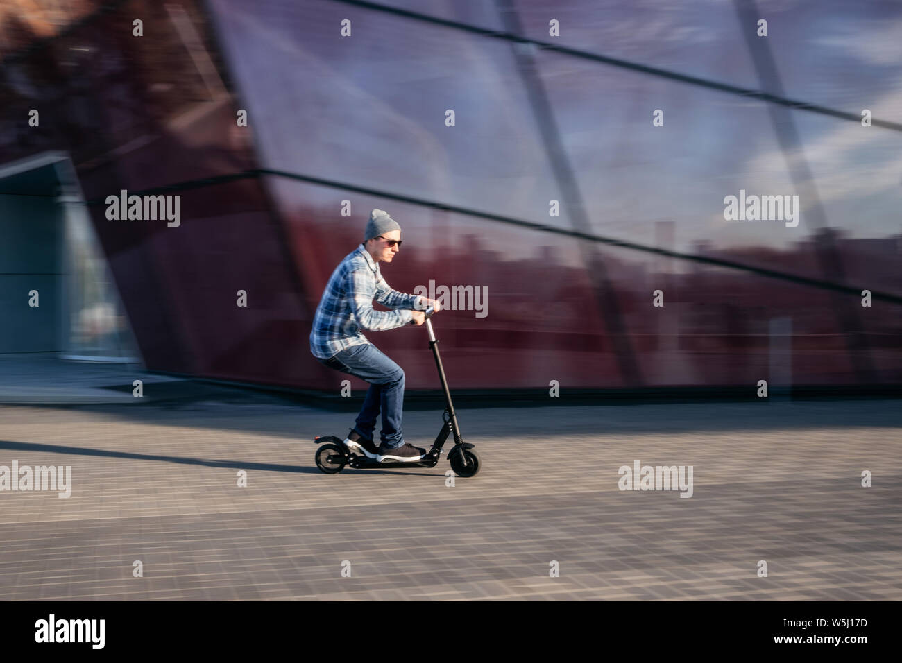 Jeune homme dans des vêtements décontractés sur electric scooter de coup on city street in motion blur contre l'immeuble de bureaux moderne en verre Banque D'Images
