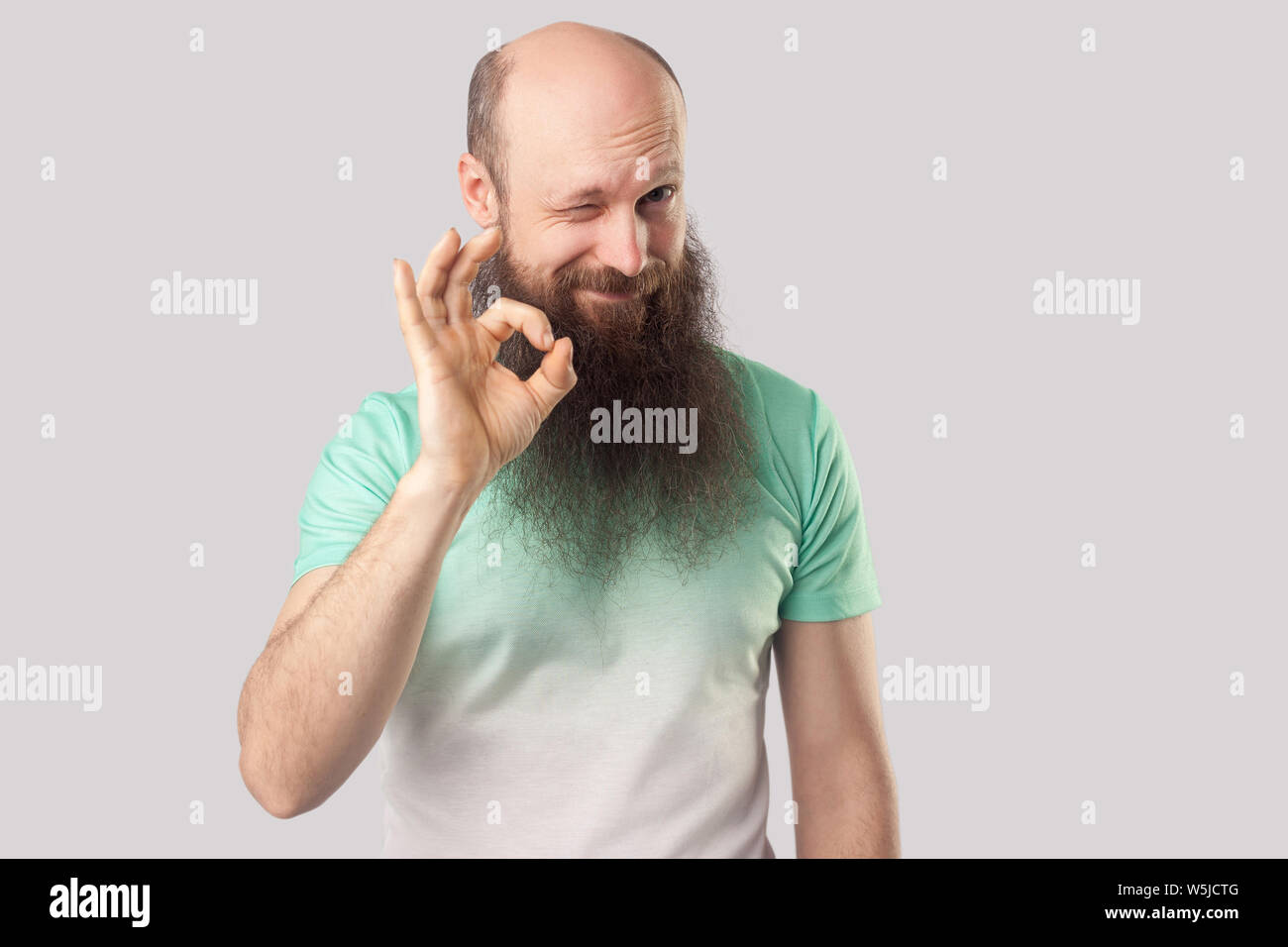 Portrait d'âge moyen drôle homme chauve avec longue barbe en vert clair t-shirt, et clignant de l'article looking at camera avec Ok sign geste. Piscine stu Banque D'Images