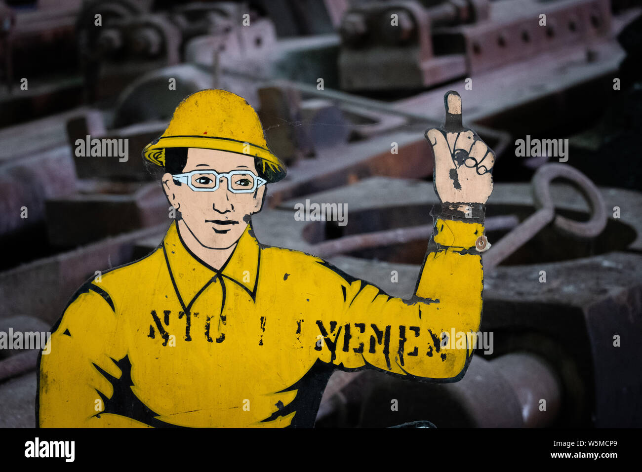 Découpe d'un personnage de ligne de dessin animé avec un casque jaune et un doigt pointant, placé sur un fond industriel Banque D'Images