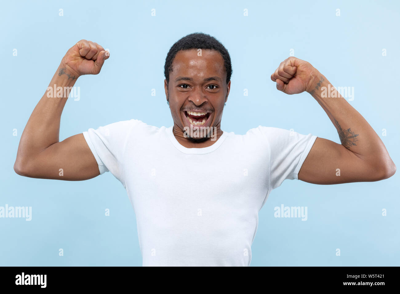 Demi-longueur close up portrait of young african-american man en chemise blanche sur fond bleu. Les émotions humaines, l'expression faciale, ad, concept. La célébration, se demande, étonné, choqué, fou heureux. Banque D'Images